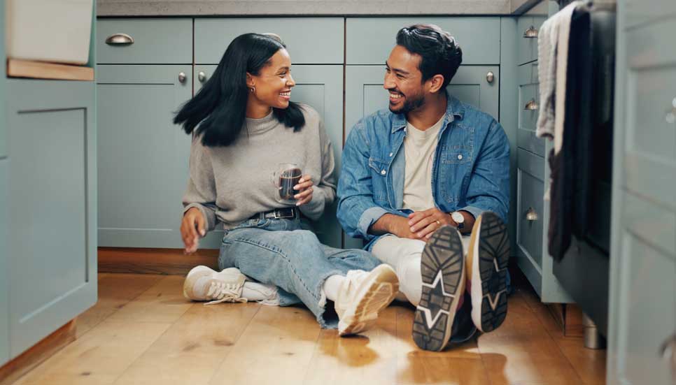 A smiling couple sits on the floor of a light teal kitchen.
